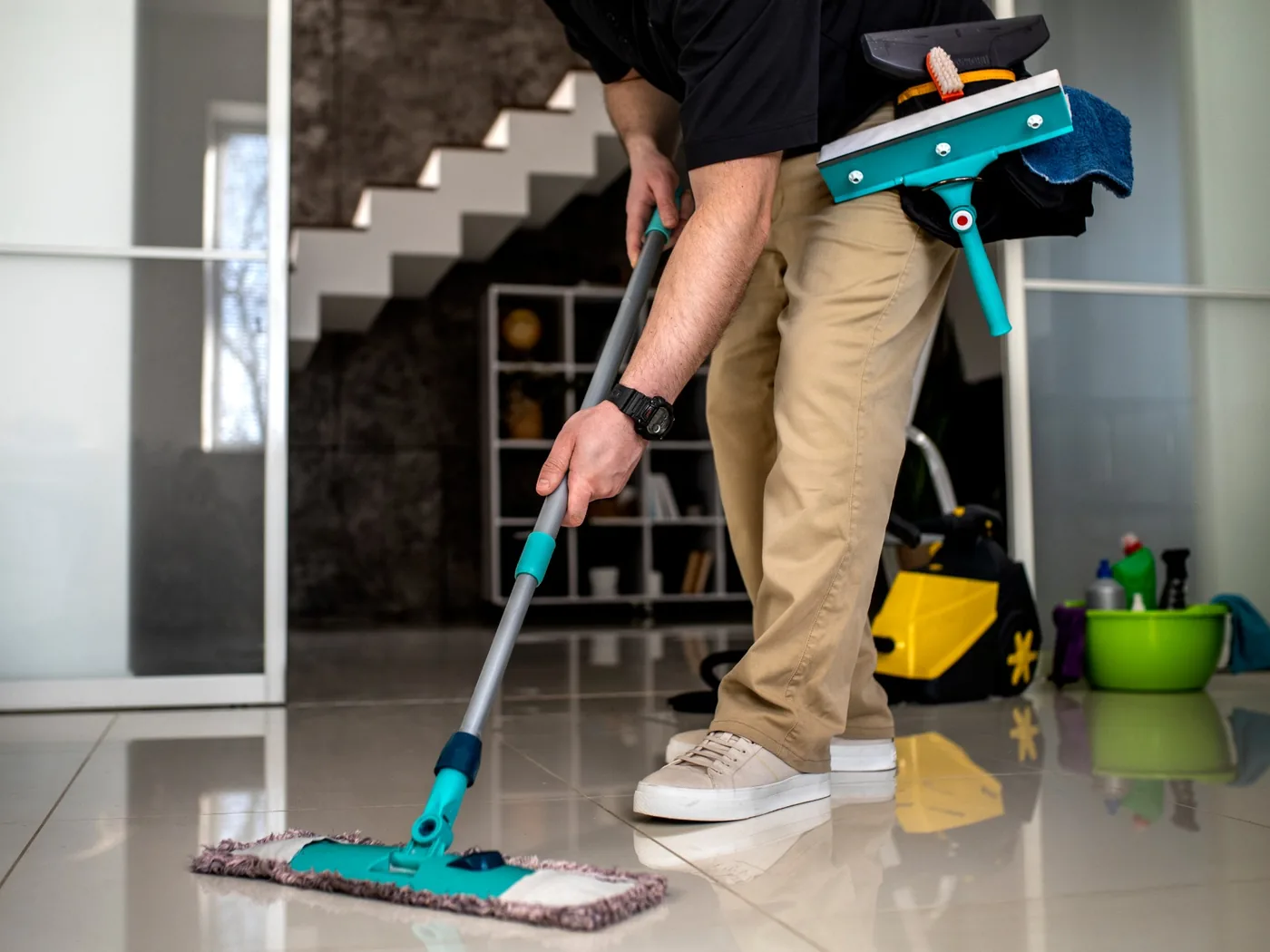 A cleaner mopping a tiled floor in a finished interior, carrying a squeegee and brush on a tool belt.