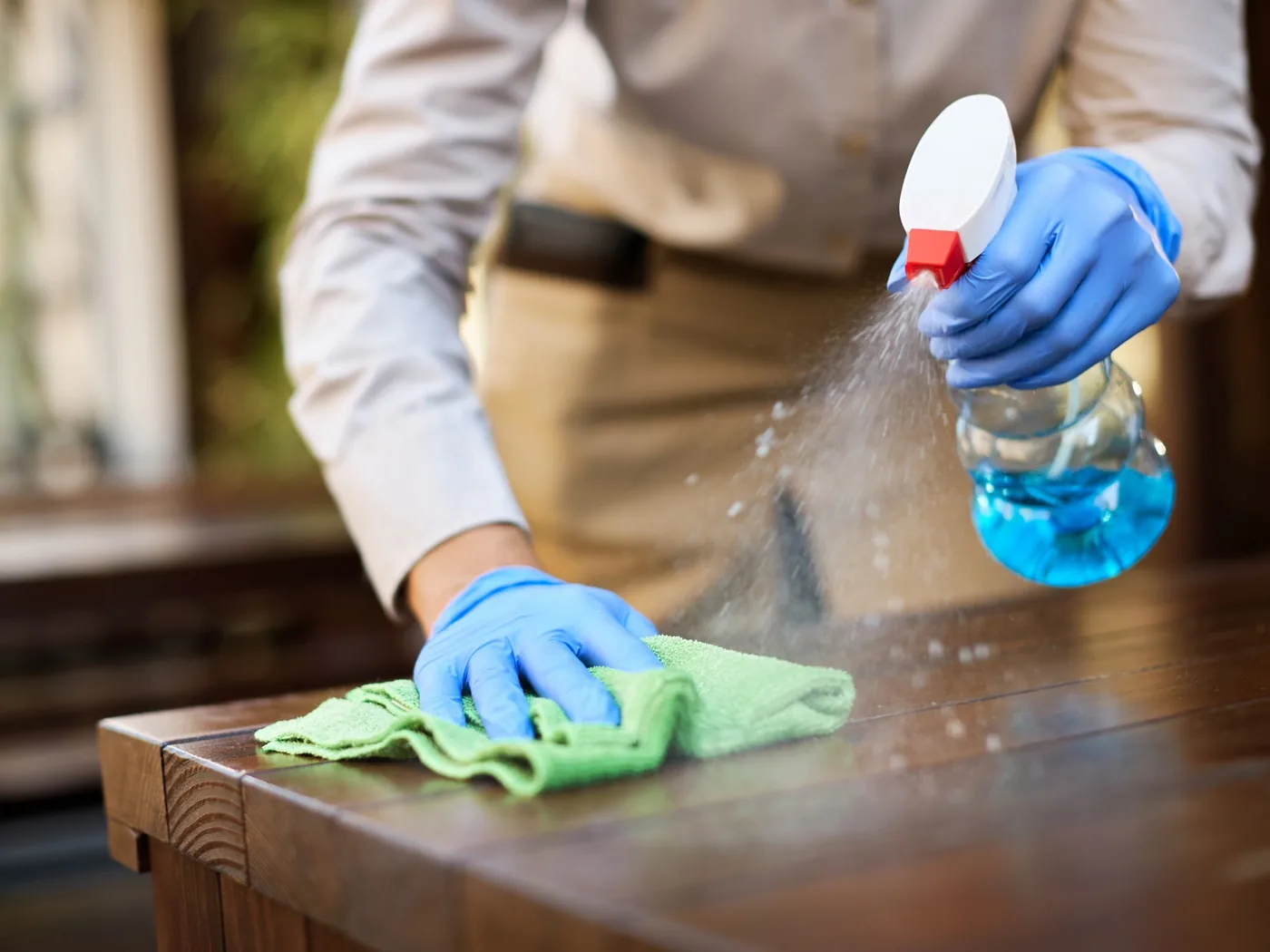 A cleaner in gloves spraying and wiping down a wooden table.